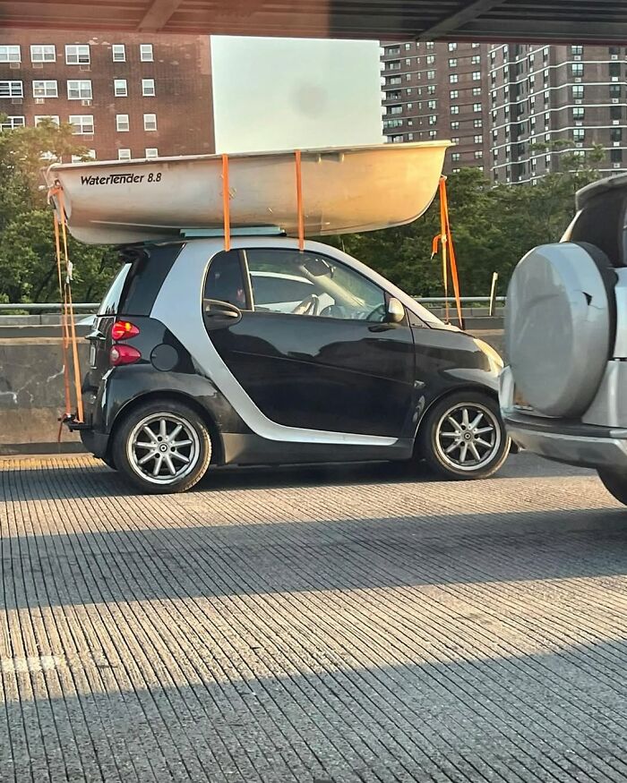 Smart car in New York with a boat strapped on top, creating a funny and unexpected urban scene.