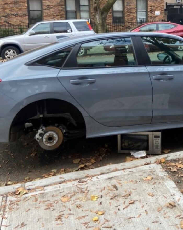 Car on New York street propped with a microwave where a wheel should be, showcasing urban humor.