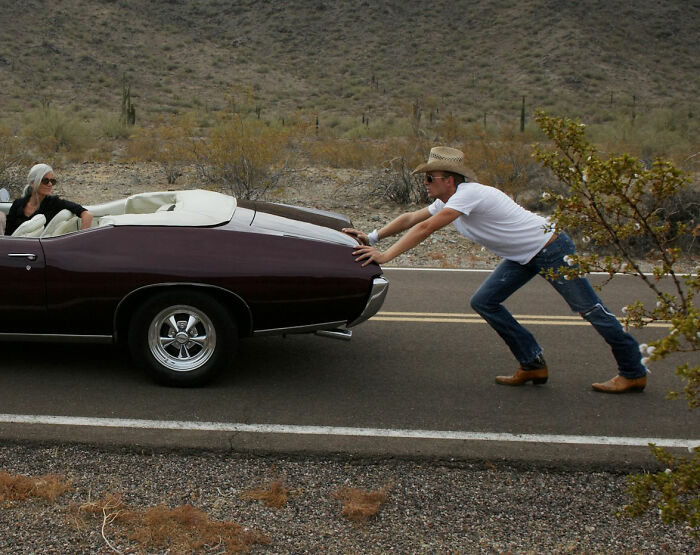 Man in cowboy hat pushing a convertible on a desert road, illustrating a moment realizing dating an idiot.