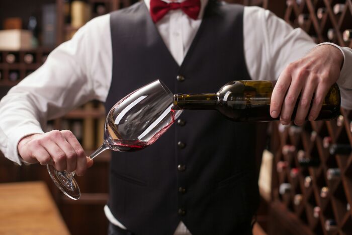 Rich kid in a wine cellar pouring red wine into a glass while wearing a formal vest and bow tie.