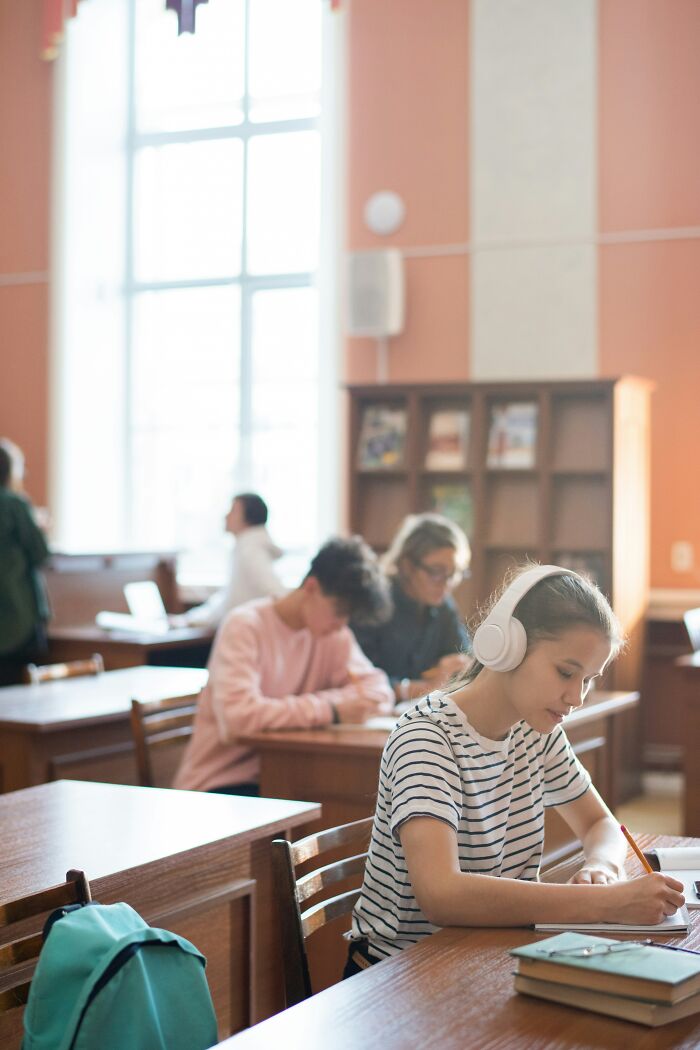 Student in a library, wearing headphones and writing, while others study quietly nearby.