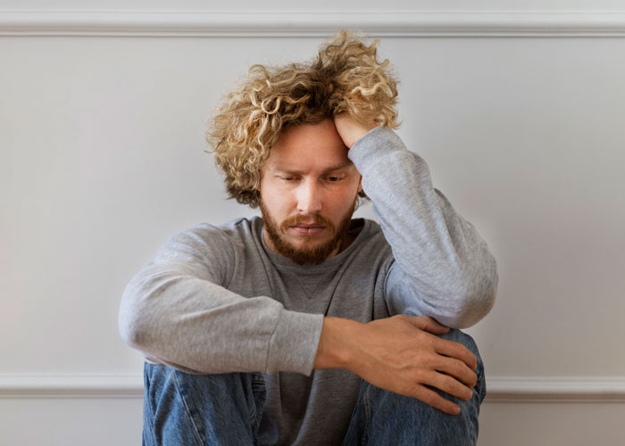 A man with curly hair, wearing a gray sweater, looking troubled, sitting on the floor