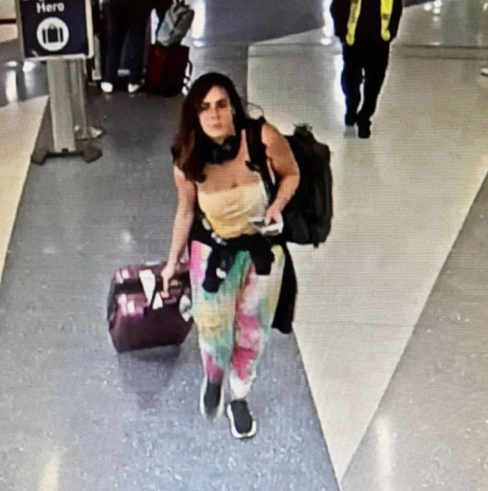 Woman at airport linked to green card marriage scam, carrying a suitcase and wearing colorful pants.