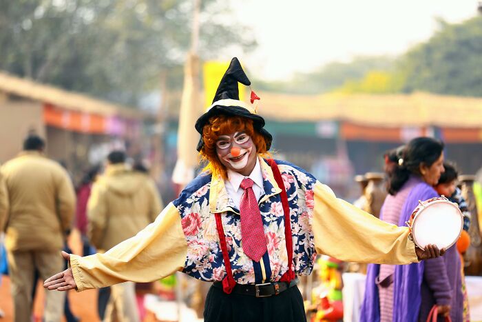 Clown smiling in colorful costume, holding a tambourine at an outdoor event, linked to professions seen as relationship red flags.