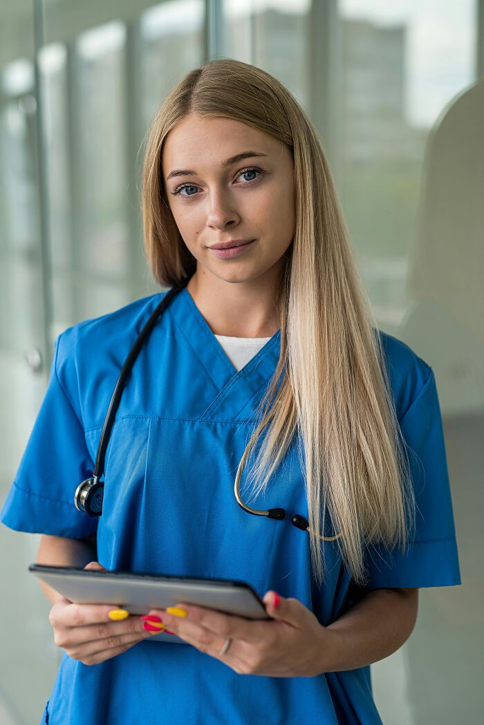 Young nurse in blue scrubs holding a tablet, standing in a bright hallway, representing professions seen as relationship red flags.