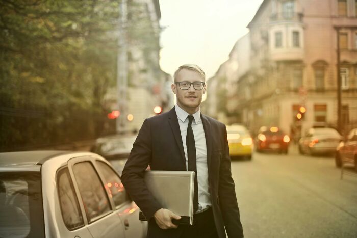 Man in a suit holding a laptop, representing a profession often considered a relationship red flag, standing on a city street.