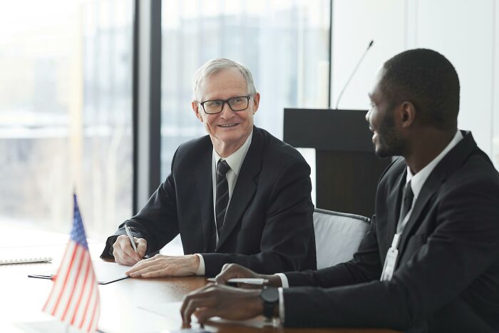 Two businessmen in suits discussing at a conference table with an American flag, related to relationship red flags in professions.