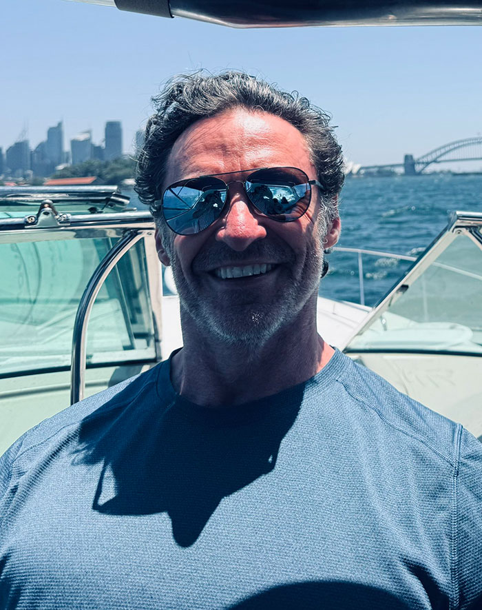 Man in sunglasses and blue shirt on a boat, with a city skyline and bridge in the background, sparking rumors.