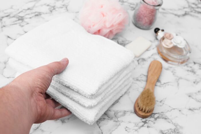 A hand placing white towels on a marble surface with frugal home essentials like a brush and pink pouf nearby.