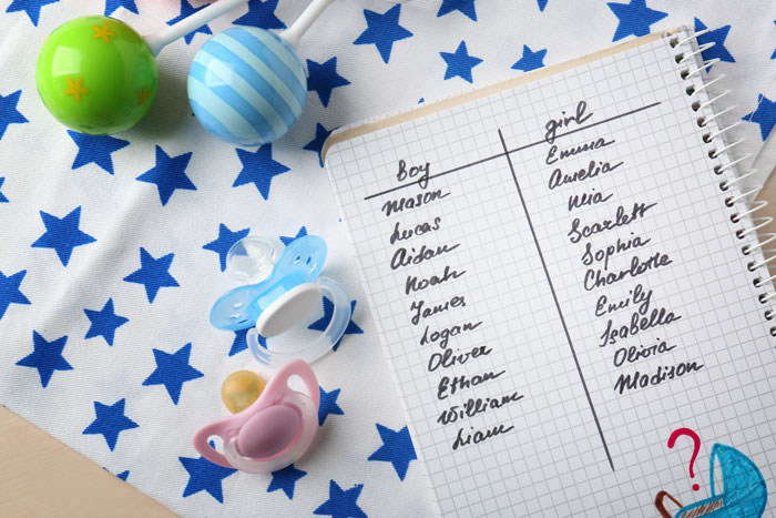 Notebook with baby names list next to pacifiers and baby toys on a star-patterned cloth.