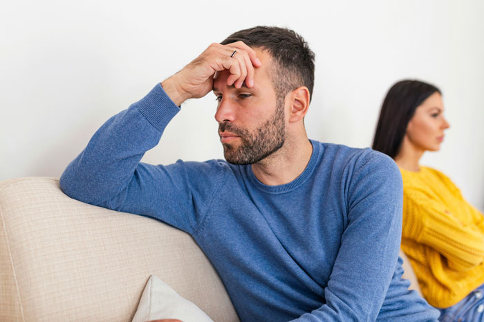 Husband in blue sweater sits on couch looking upset; wife in yellow sweater sits in background, symbolizing name veto conflict.