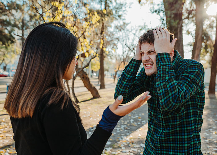 A couple arguing in a park, with the husband looking defensive, wearing a green plaid shirt.