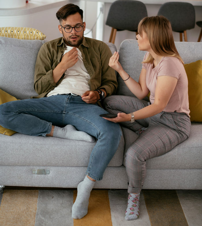 Couple discussing Hawaii trip on sofa, woman upset, man suggesting after multiple Disney visits.