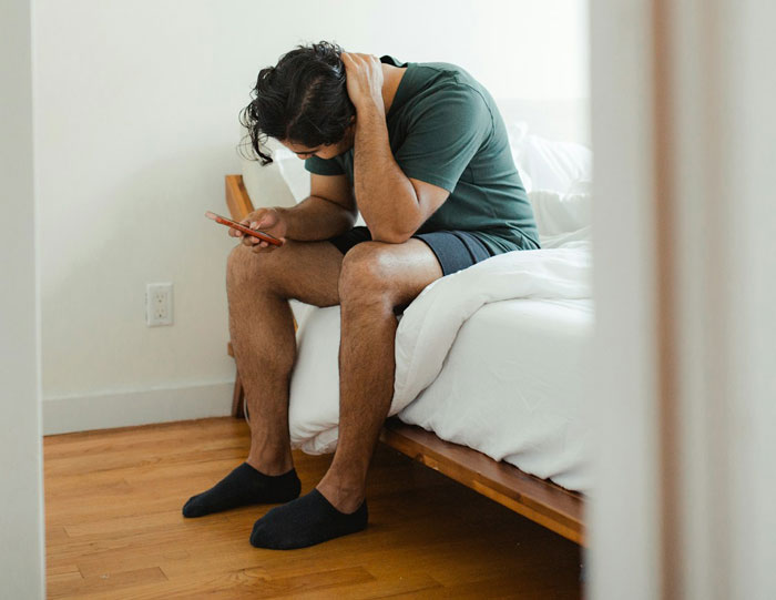 Man in casual attire sitting on a bed, looking at phone, contemplating vacation change from Disney to Hawaii.