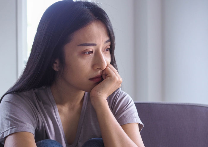 A woman with long hair looks thoughtful and pensive, wearing a gray shirt, inside a room.