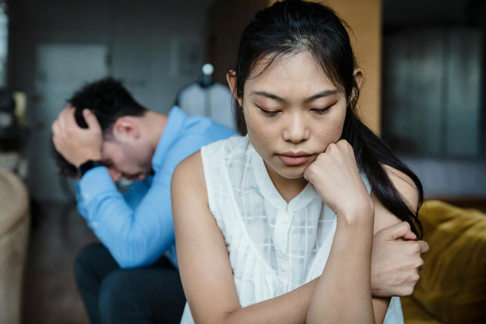 A distressed couple sits separately; focus on wife reflecting on Asian features.