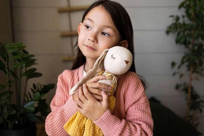 Child with Asian features holding a stuffed doll, wearing a pink sweater, looking pensive.