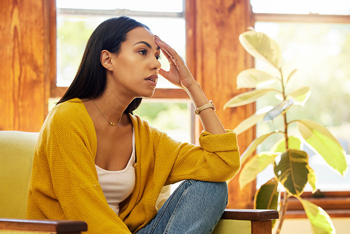 Woman feeling upset about husband's "work wife," sitting by window, wearing yellow cardigan.