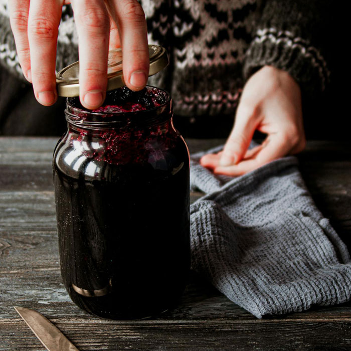 Woman's hands sealing a jar of jam on a wooden table, with a grey cloth beside it.