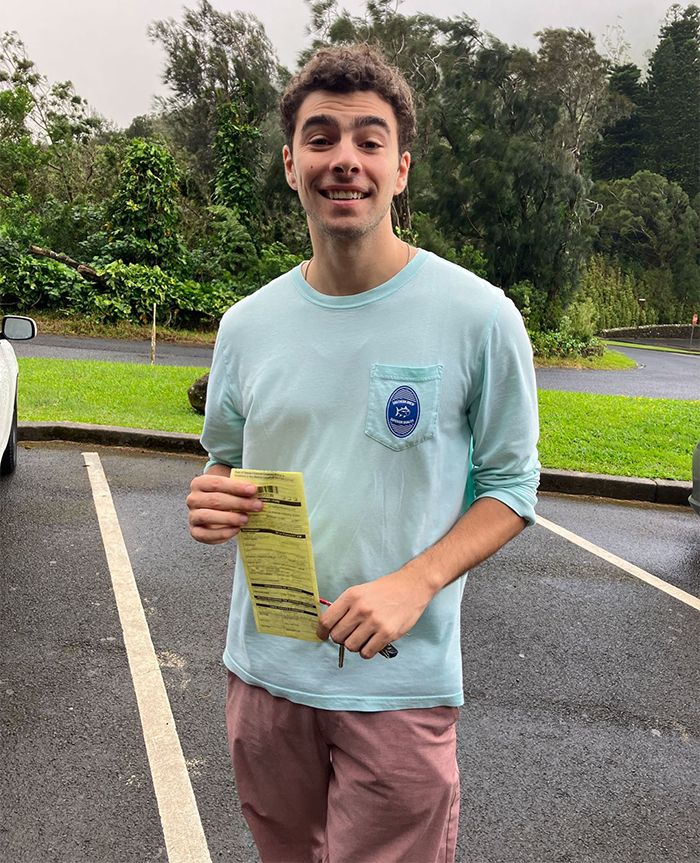 Luigi Mangione in a light blue shirt holding a paper in a parking lot, surrounded by greenery.