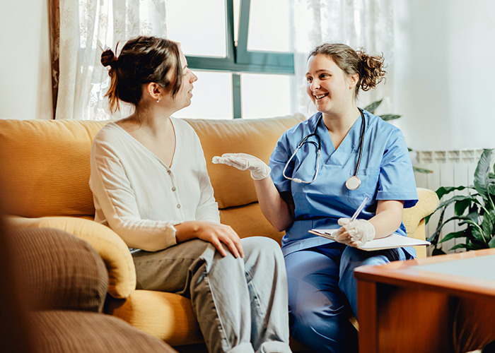 Nurse in blue scrubs discussing a report with a woman on a sofa, related to insurance horror stories.