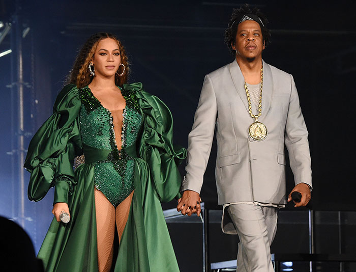 Jay-Z on stage wearing a gray suit, walking and holding hands with wife Beyonce, amid recent lawsuit allegations.