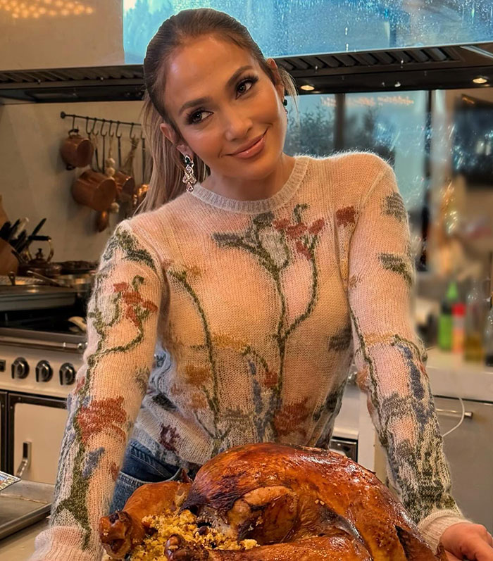 Jennifer Lopez in a kitchen with a floral sweater, leaning on a counter with a roasted turkey.