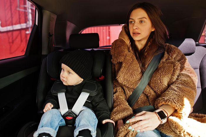 A woman appearing upset while sitting beside a child in a car, wearing a brown coat.