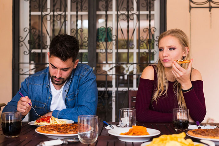 A couple at a dinner table, the woman appears upset, while the man focuses on his meal; theme of joke accident pregnancy.