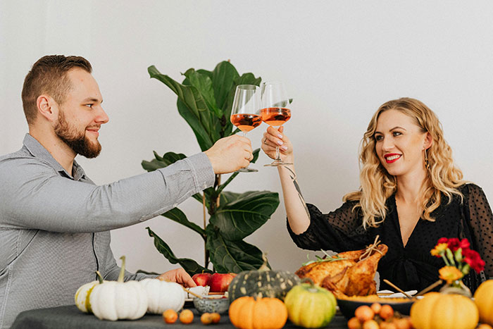 A couple toasting with wine at a festive dinner, surrounded by pumpkins and a turkey, smiling brightly.