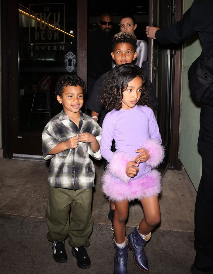 Kanye West and a woman associated with his new wife are seen in the background, with children dressed stylishly in the foreground exiting a venue.