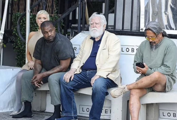 Kanye West sitting on a bench with a woman associated with his new wife, alongside two older men, one on his phone, in a casual outdoor setting.