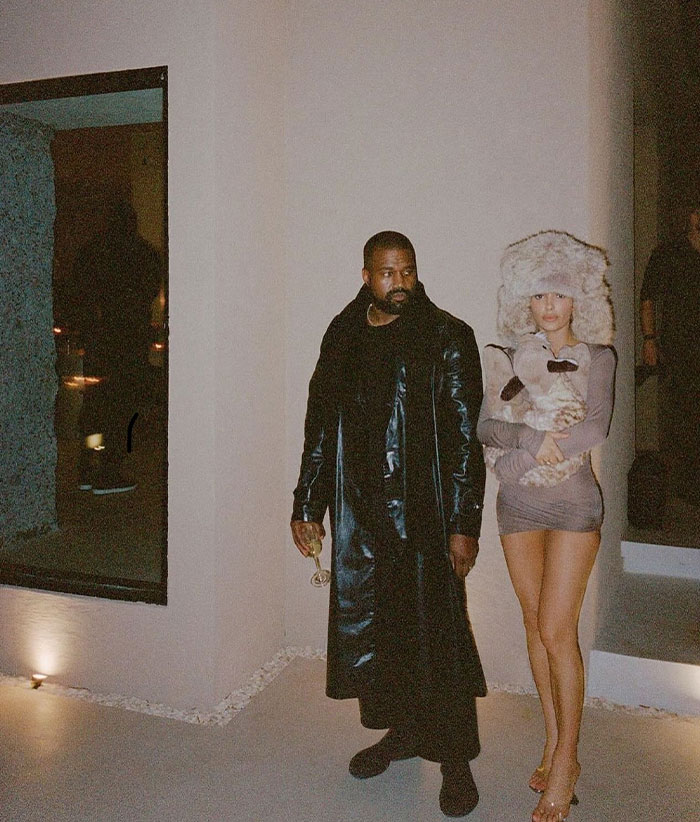 Kanye West in a black leather coat standing beside a woman associated with his new wife, wearing a furry hat and a short dress, in an elegant setting.