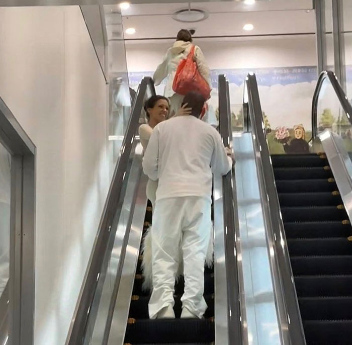 Kanye West dressed in white on an escalator, facing and holding a smiling woman associated with his new wife, in a bright indoor setting.