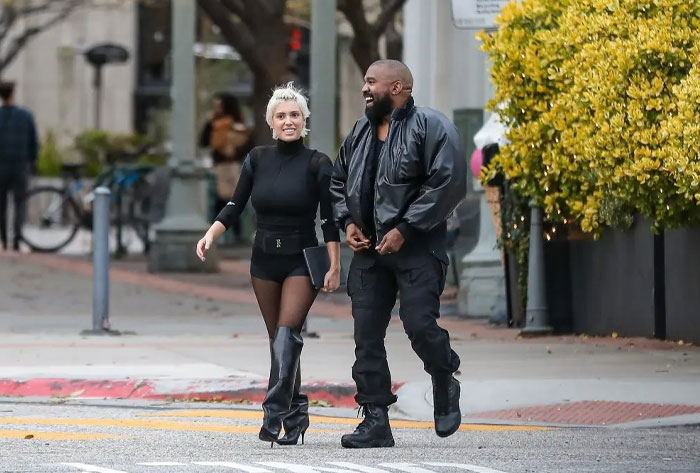 Kanye West in a black outfit walking and smiling with a woman associated with his new wife, dressed in a stylish black ensemble, on a city street.