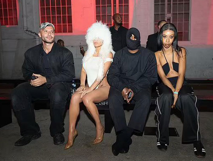 Kanye West in a masked black outfit sitting beside a woman associated with his new wife, wearing a furry white hat and minimalist attire, at an event with two others.