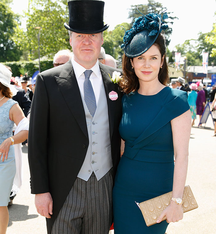 A couple in formal attire at an outdoor event, with the man wearing a top hat and the woman in a teal dress and matching hat.