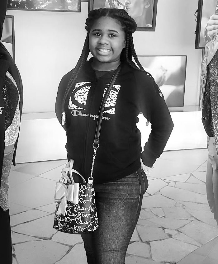 Kelaia Turner smiling in a casual setting, wearing a Champion hoodie and carrying a patterned bag, related to bullying and abuse.