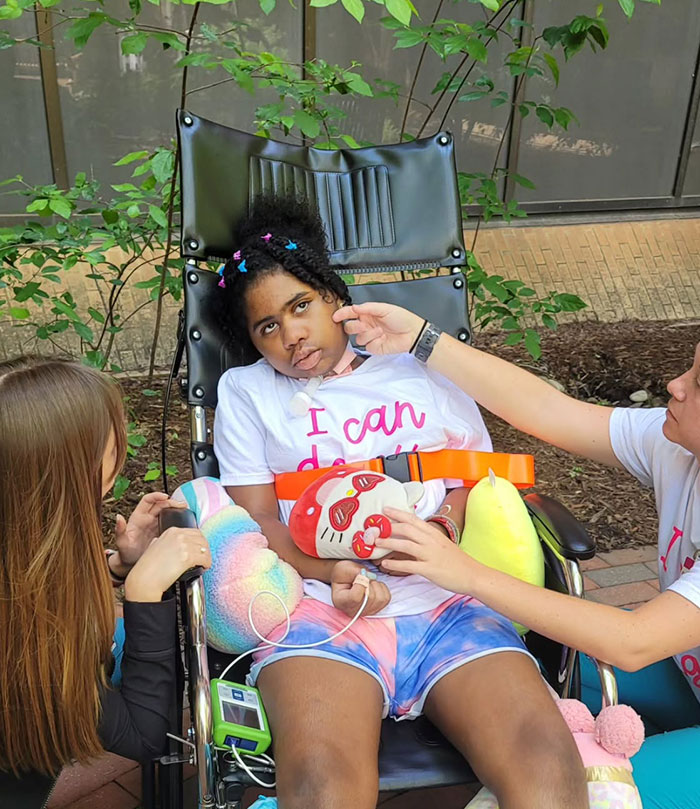 Kelaia Turner in a wheelchair outside, surrounded by supportive friends, holding plush toys.