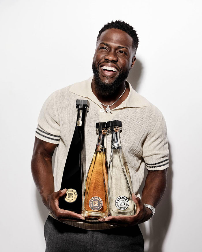 Kevin Hart smiles broadly while holding three bottles of tequila from his Gran Coramino brand, wearing a casual collared sweater.