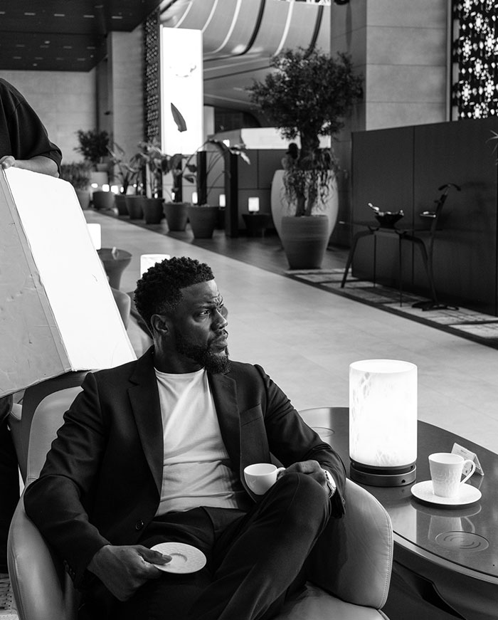 Kevin Hart sits in a modern lounge setting, holding a teacup and saucer, wearing a sleek suit, looking off thoughtfully in a black-and-white image.