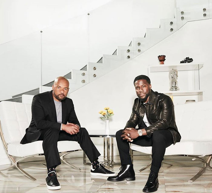 Kevin Hart sits with a business partner in a sleek, modern interior featuring white furniture, a glass staircase, and minimalist decor.