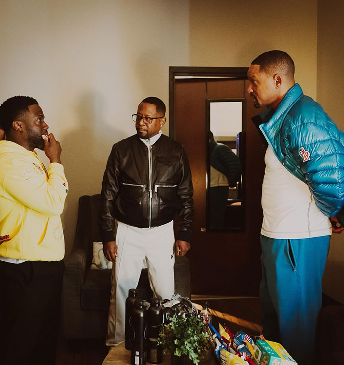 Kevin Hart in a yellow sweater conversing with two men, one in a black leather jacket and another in a blue puffer jacket, in a casual setting.