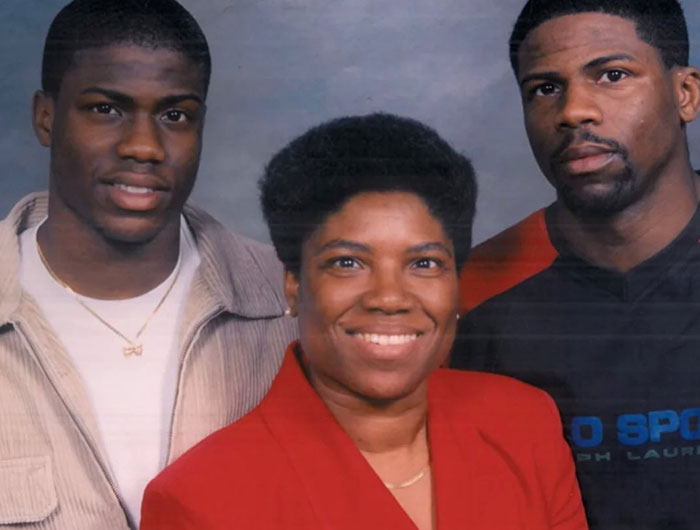 A younger Kevin Hart poses with his mother and brother in a family portrait, all smiling warmly against a studio backdrop.