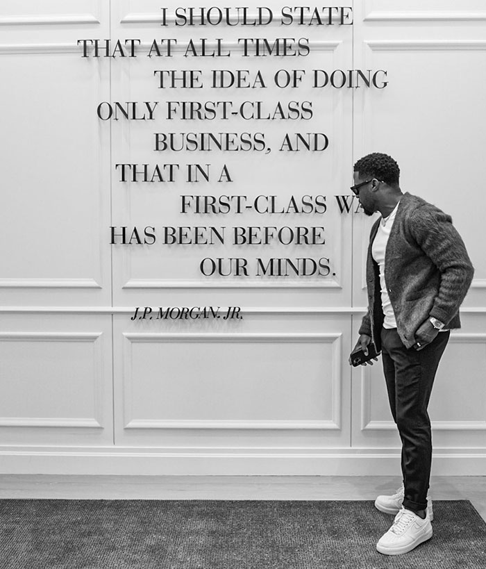 Kevin Hart observes a wall inscription by J.P. Morgan Jr. about first-class business, dressed casually in a cardigan and sneakers.