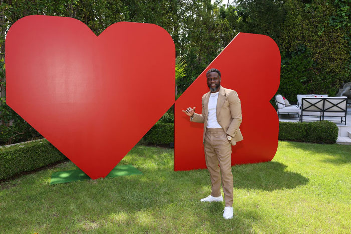 Kevin Hart poses outdoors in front of a large red heart sculpture, wearing a tan suit and white sneakers, smiling confidently.