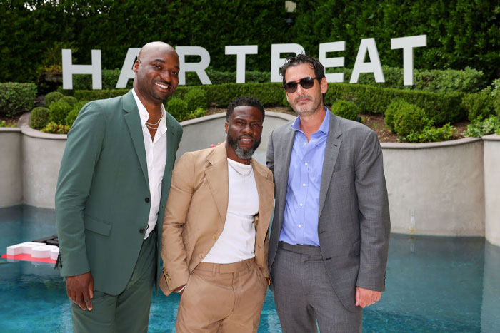 Kevin Hart poses with two men in formal attire by a pool, with the "Hartbeat" sign visible in the background, all smiling.