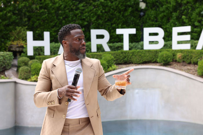 Kevin Hart speaks while holding a microphone and a drink, dressed in a tan suit, with the "Hartbeat" sign visible in the background.