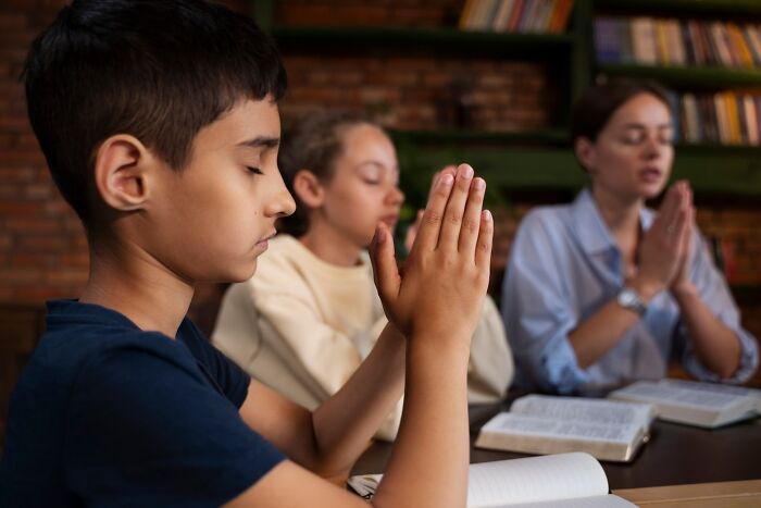 Children praying in a classroom setting, focusing intently, with books open on the table.