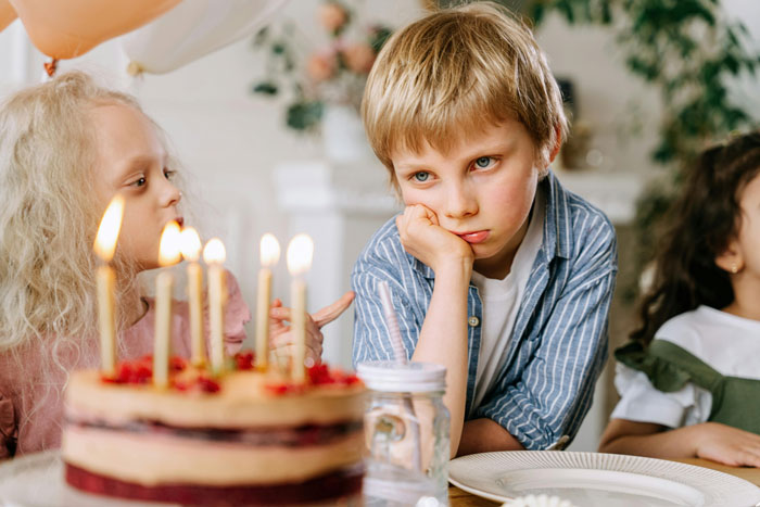 Kids at birthday party, one looking bored, near cake with lit candles.